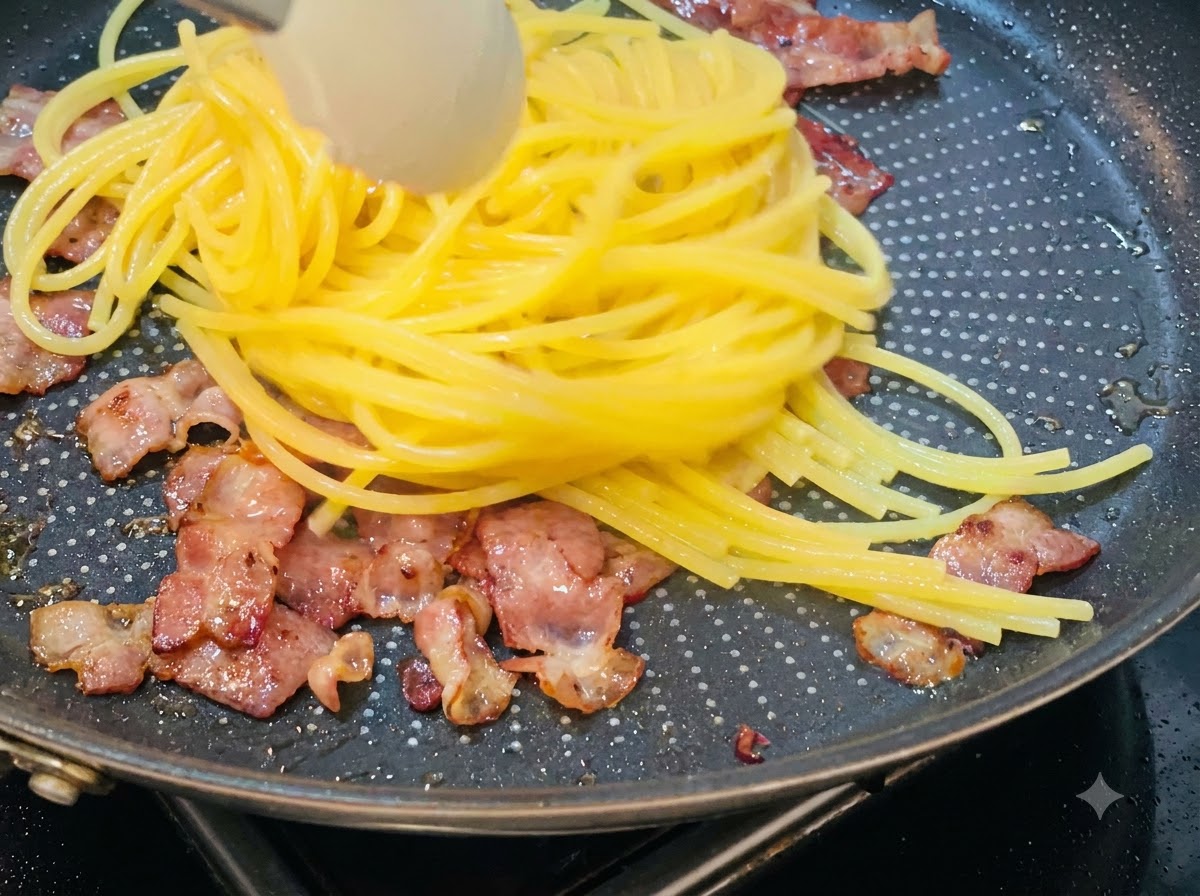 茹で上がったスパゲッティをトングで持ち上げ、カリカリのベーコンが入ったフライパンに移している様子