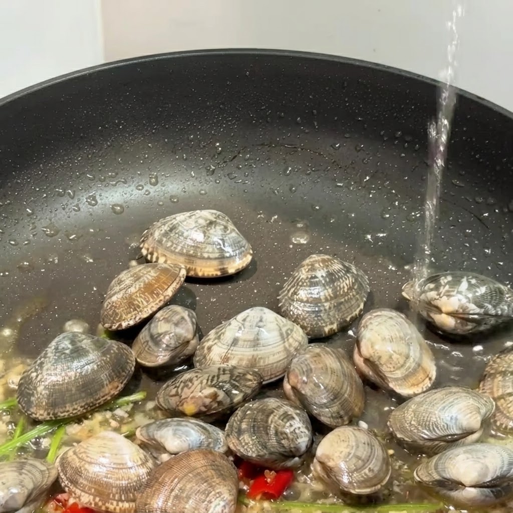 アサリ、ニンニク、赤唐辛子が入った黒いスキレットに液体が注がれ、蓋が一部見えている様子。