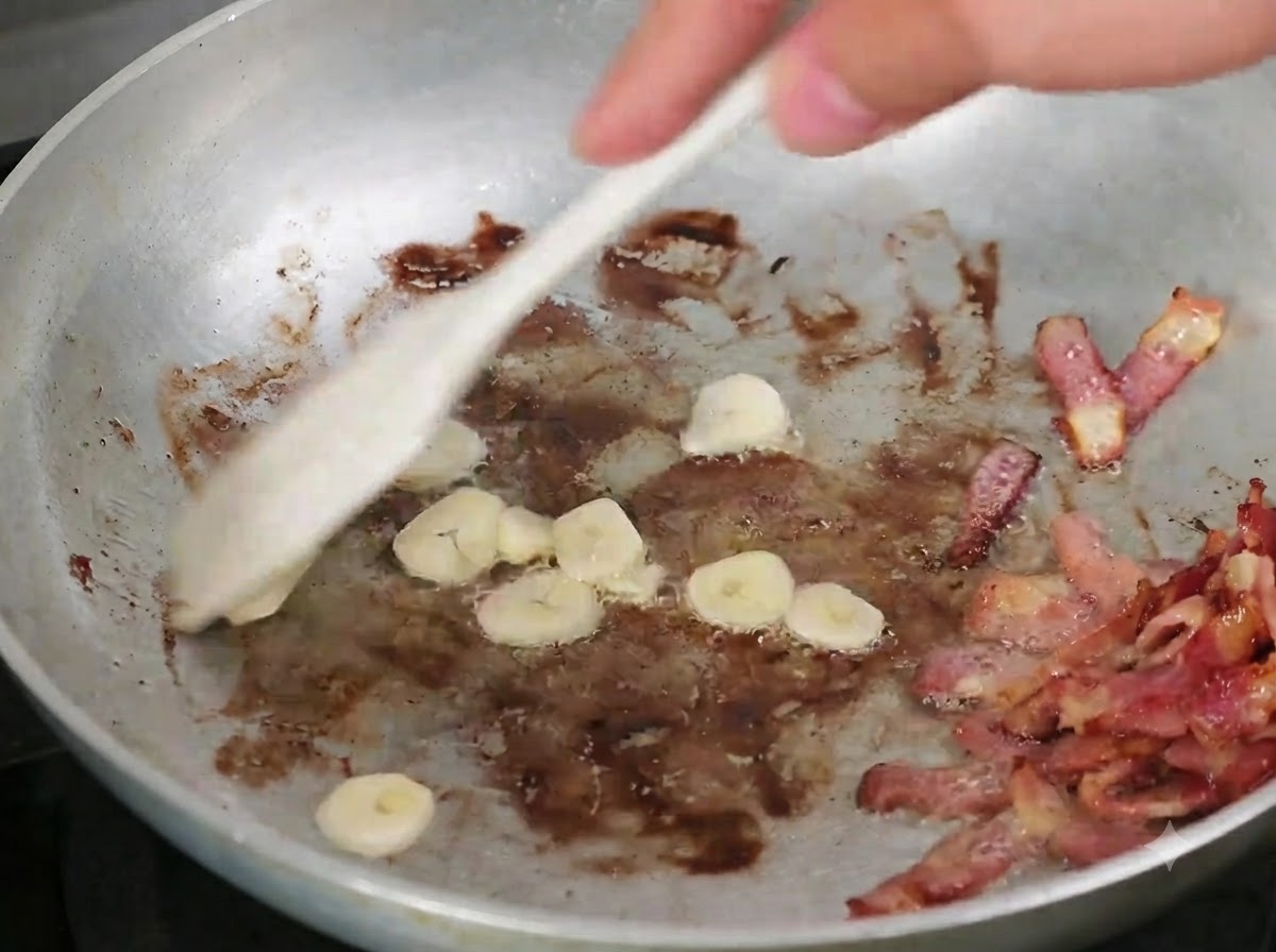 木べらを使って、炒めたベーコンが入ったフライパンにスライスにんにくが混ぜ合わされている様子。