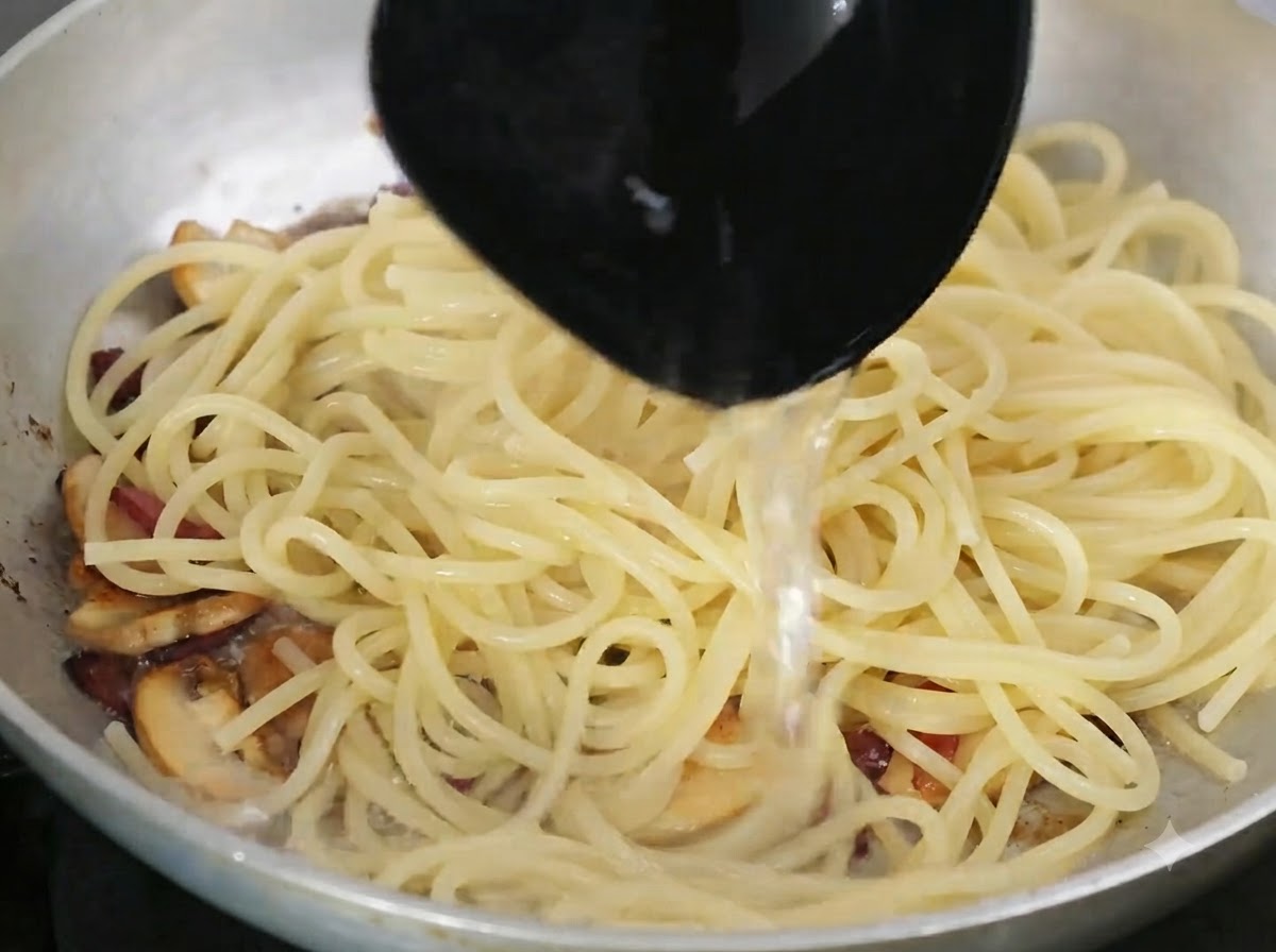 お玉でデンプン質を含んだパスタの茹で汁が、パスタと具材の入るフライパンに注がれているところ。