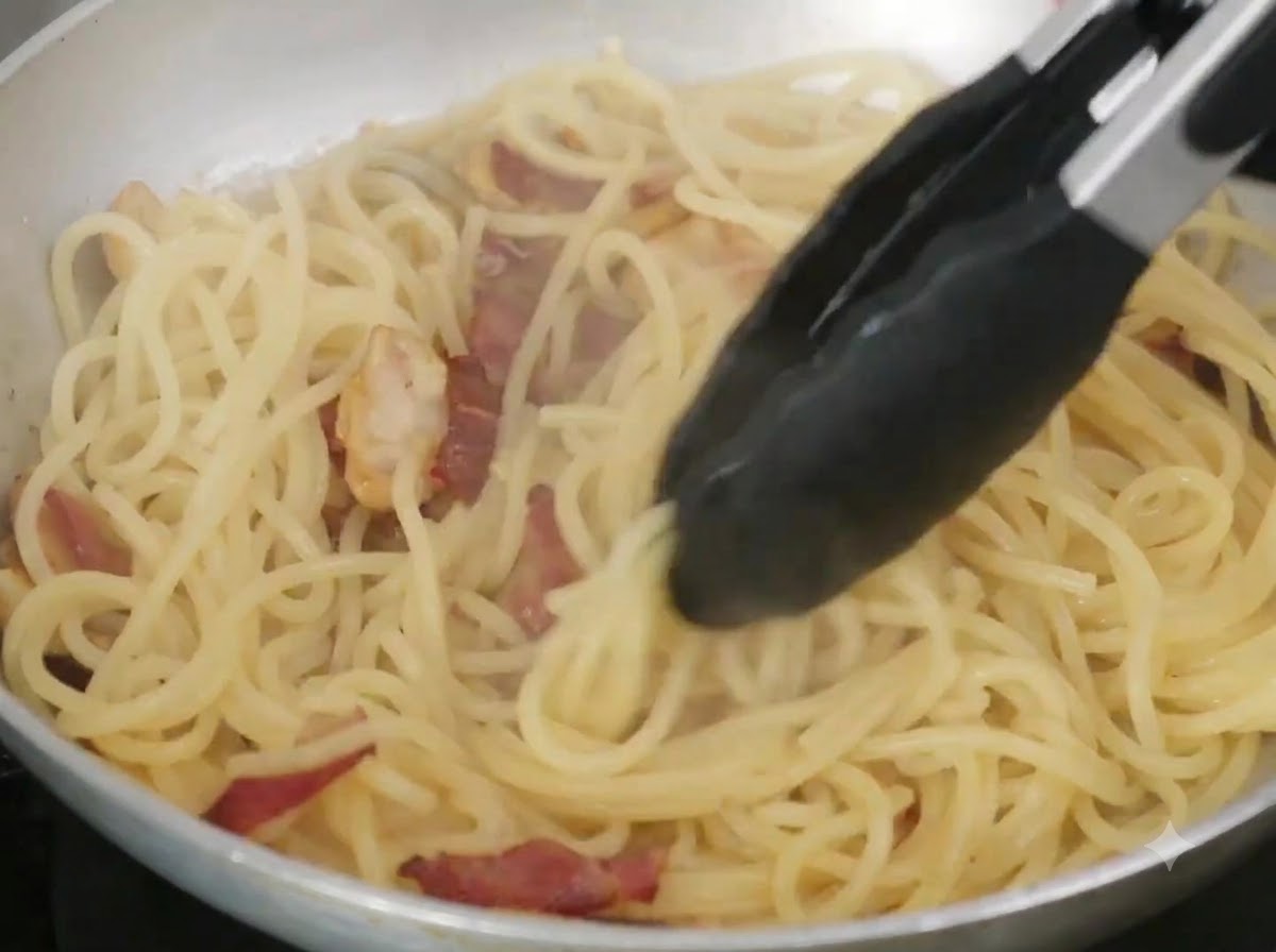 フライパンの中でトングを使い、バターとパスタの茹で汁をスパゲッティにしっかりと絡めている様子。