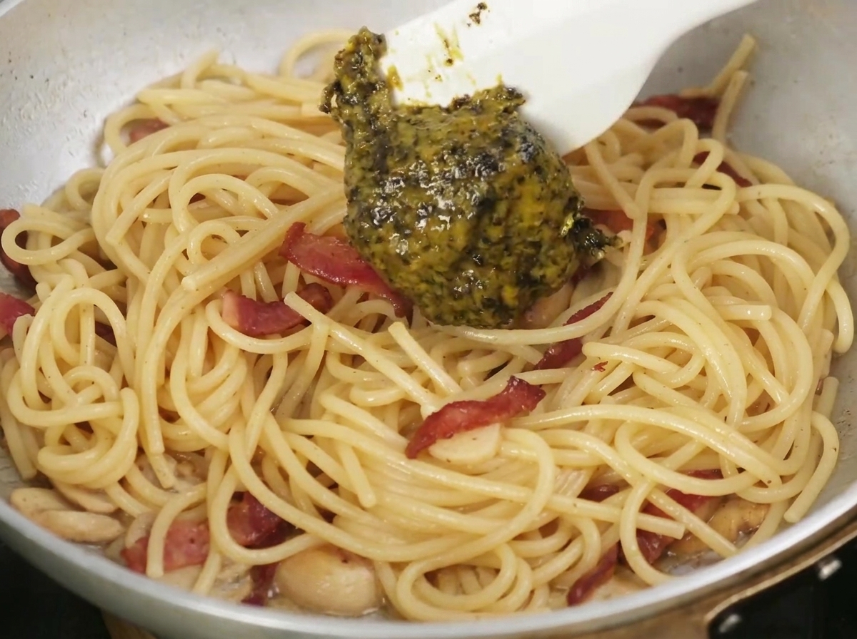 調理中のパスタとベーコンが入ったフライパンに、黒トリュフソースがスプーンで加えられているところ。