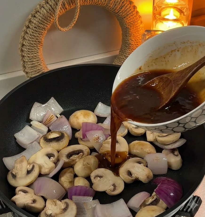 スライスした玉ねぎとホワイトマッシュルームが入ったフライパンに、ボウルに入った茶色のソースを注いでいる様子。