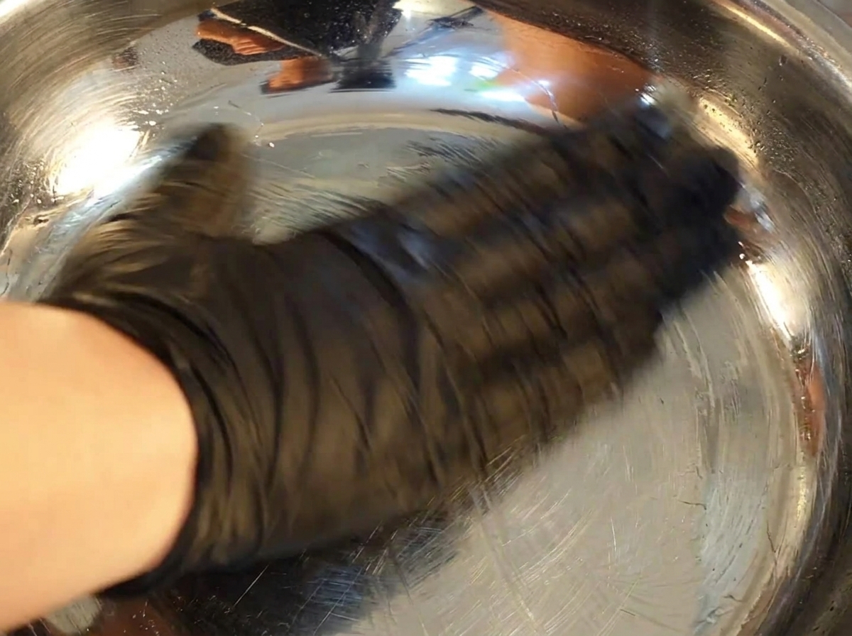黒い手袋を着用した手が、ペーパータオルを使って光沢のあるステンレスボウルの内側に油を塗っている様子。