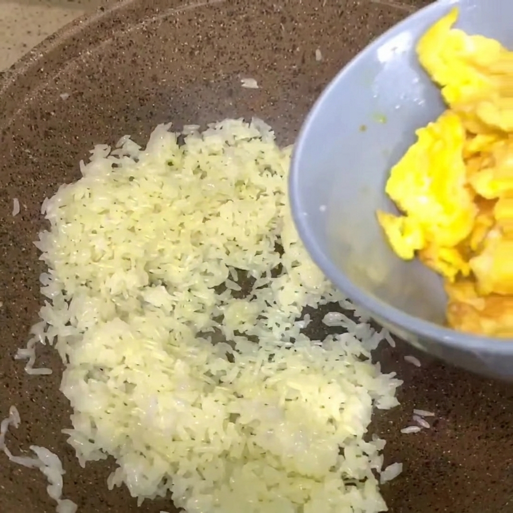 灰色のボウルに入った炒り卵が、白ご飯の入ったフライパンに投入されている様子。