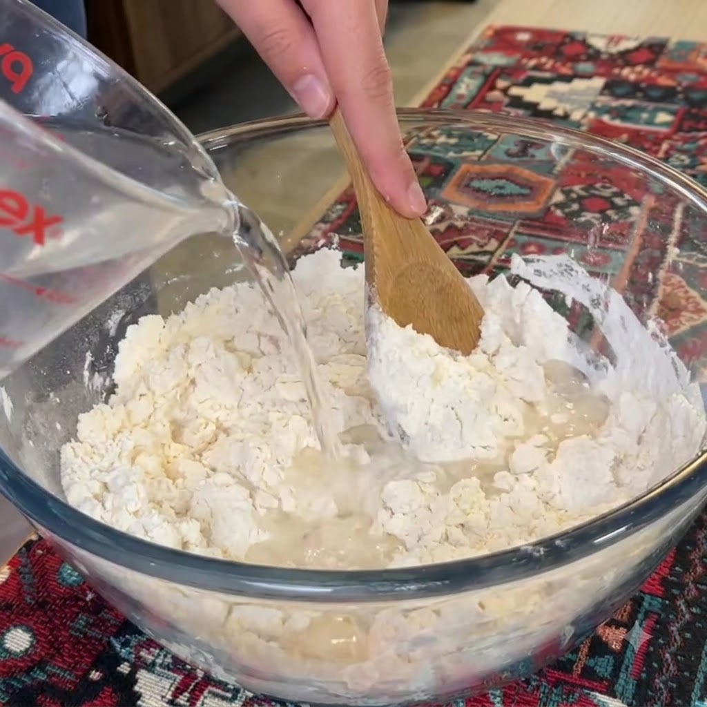木べらで小麦粉を混ぜながら、計量カップからガラスボウルへ水を注いでいる様子。