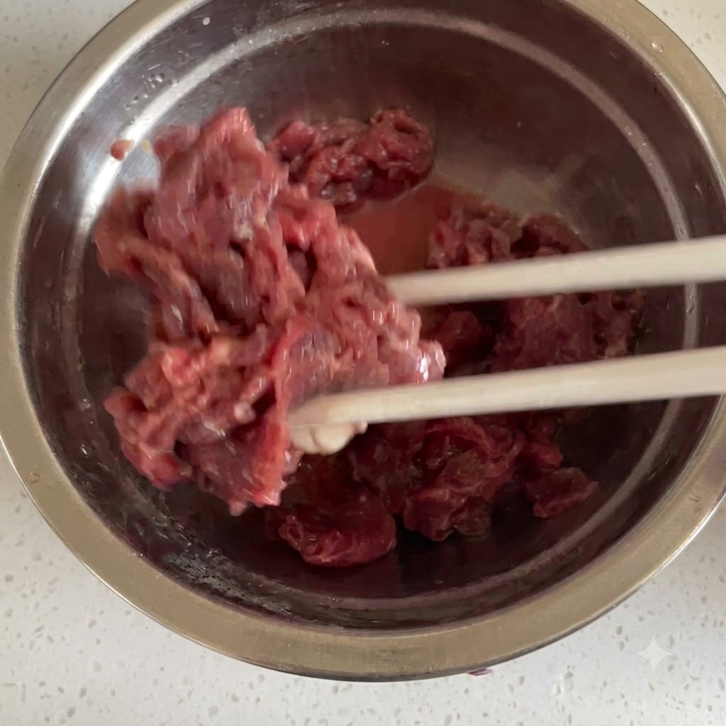 ステンレス製のボウルの中で、箸を使って薄切り生肉とマリネの材料をしっかりと混ぜ合わせている様子。
