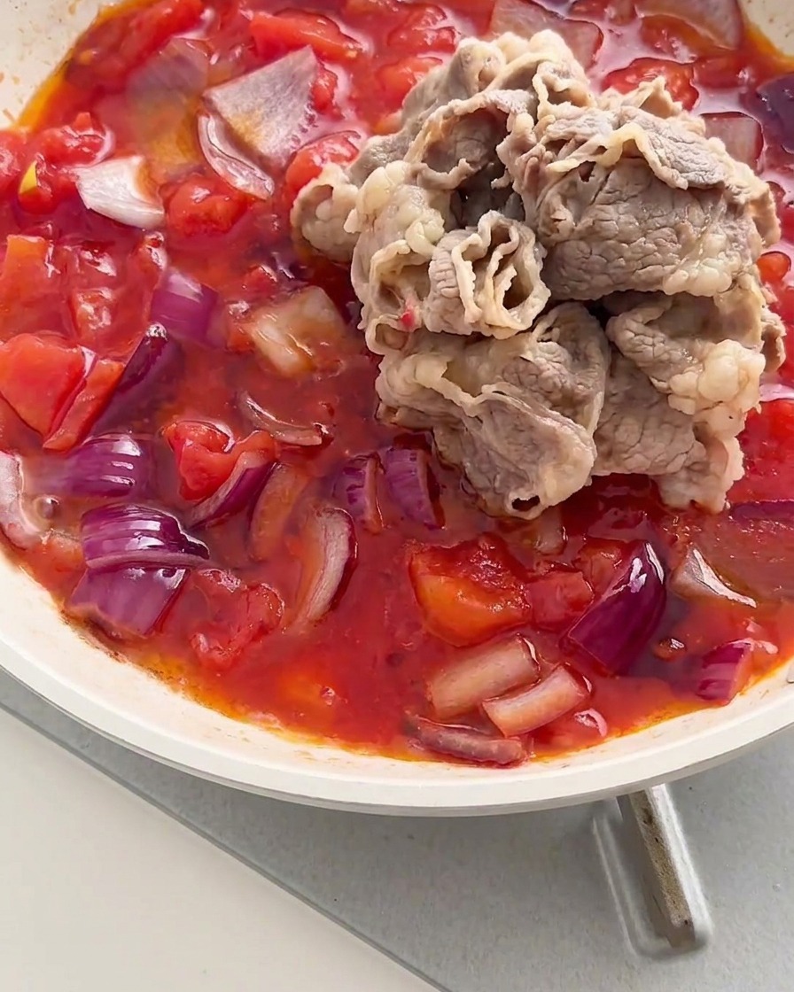 赤トマトと玉ねぎの煮込みソースの中央に、下茹でした牛肉ロールの山を入れているところ。