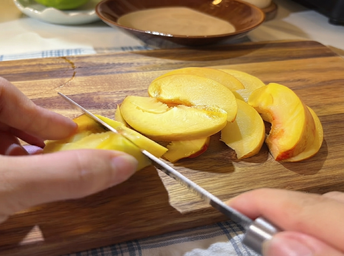 木製のまな板の上で、小さなナイフを使って新鮮な黄桃の半分を薄切りにしている手。