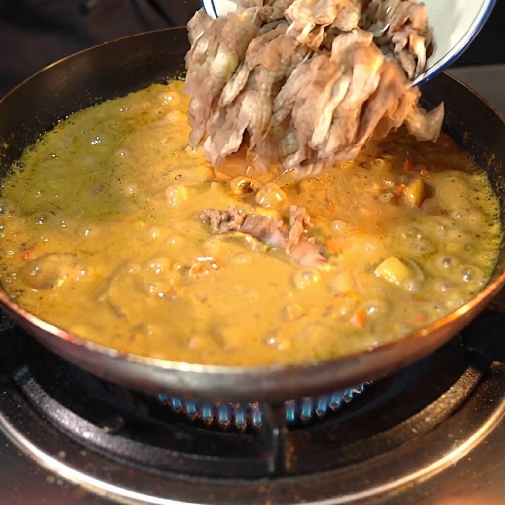 白いボウルから、濃くなった黄色いカレーソースと野菜の入った暗い色のフライパンへ注がれる下処理済みの牛肉スライス。