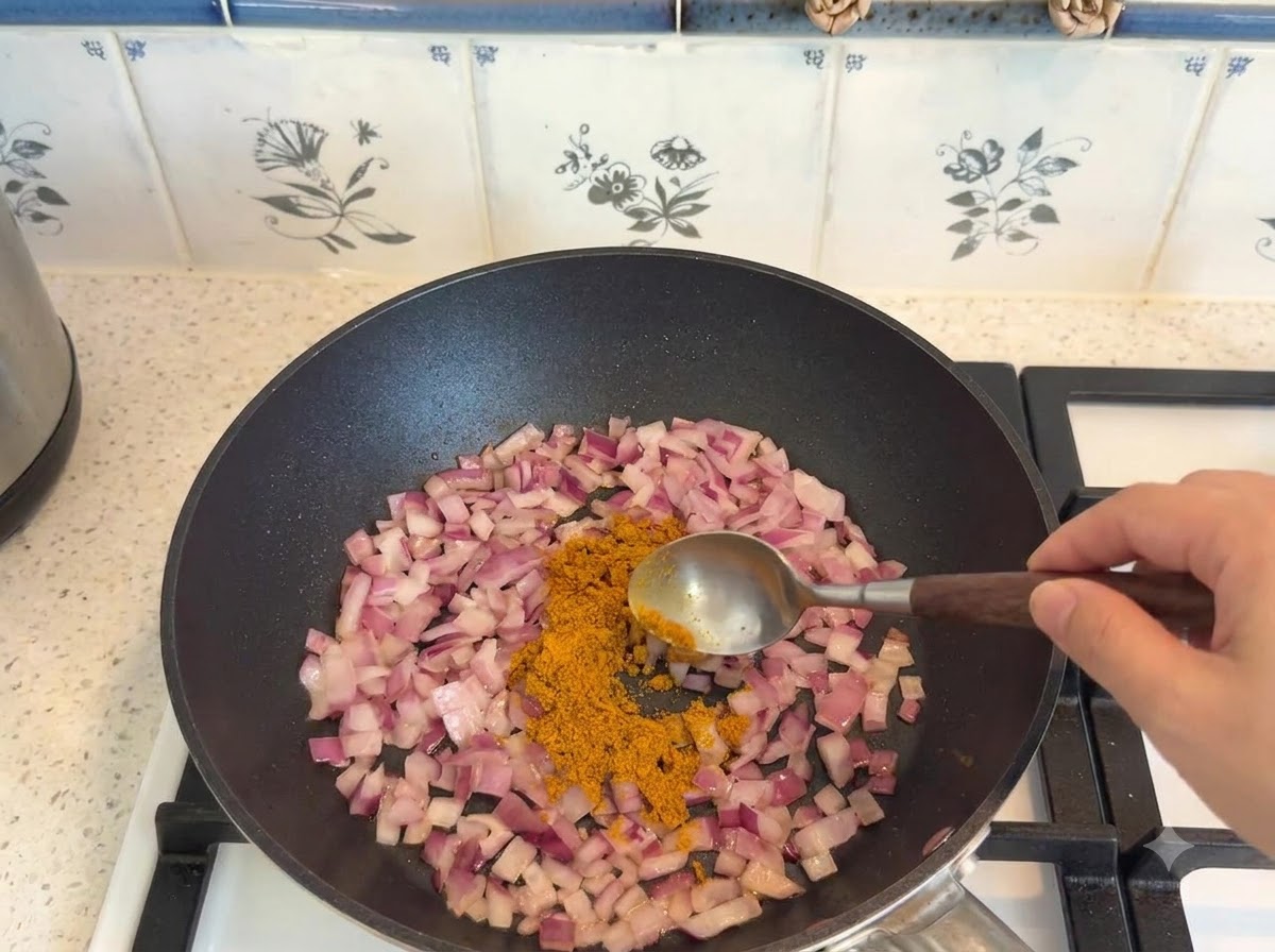 スプーンで計量されたカレー粉が、フライパンで炒めた玉ねぎに加えられている様子。