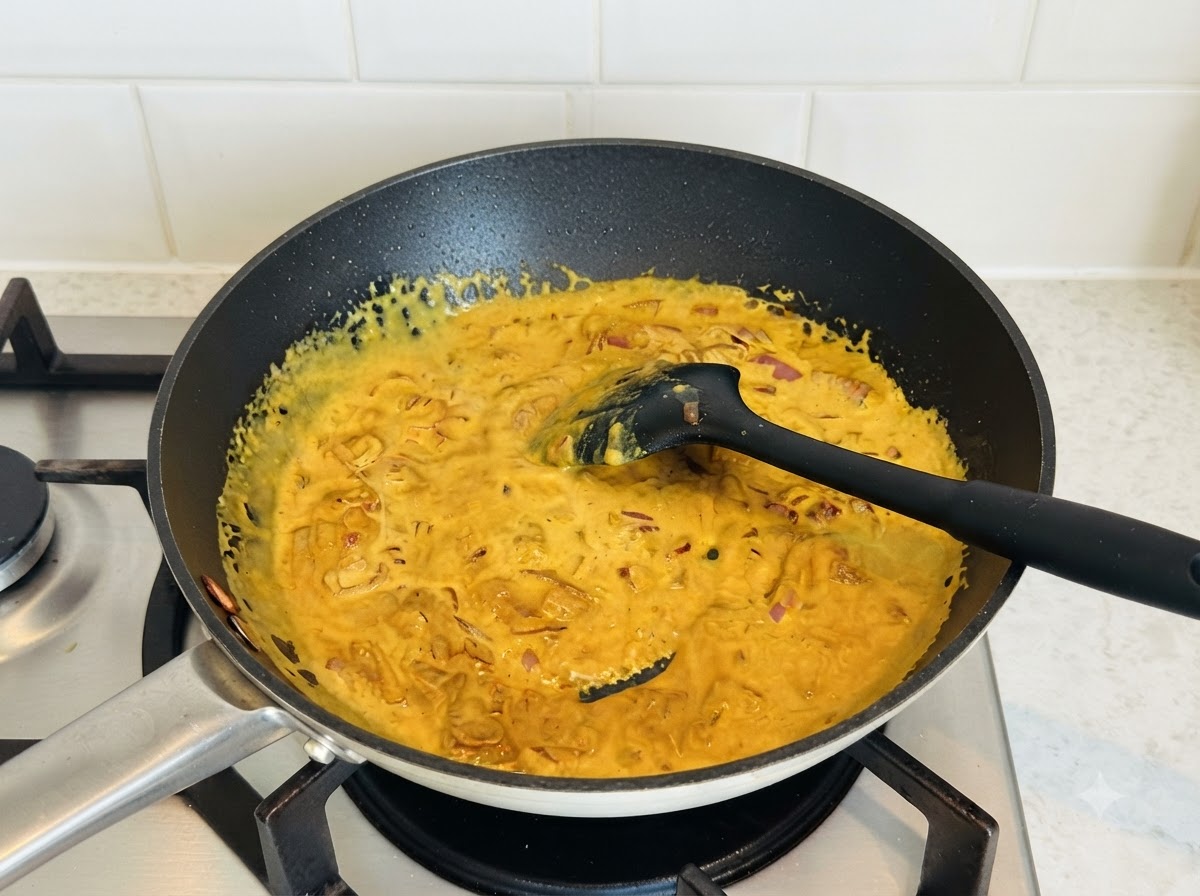 黒いシリコンスパチュラで、濃い色のスキレットの中で濃厚な黄色いカレーソースがかき混ぜられている様子。