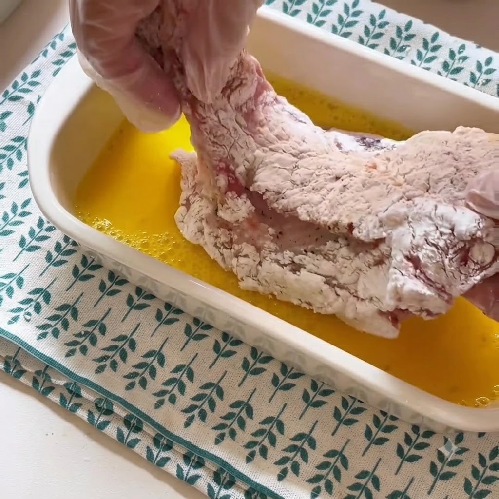 手袋をした手が、片栗粉をまぶした豚肉を黄色の溶き卵の容器から持ち上げている。