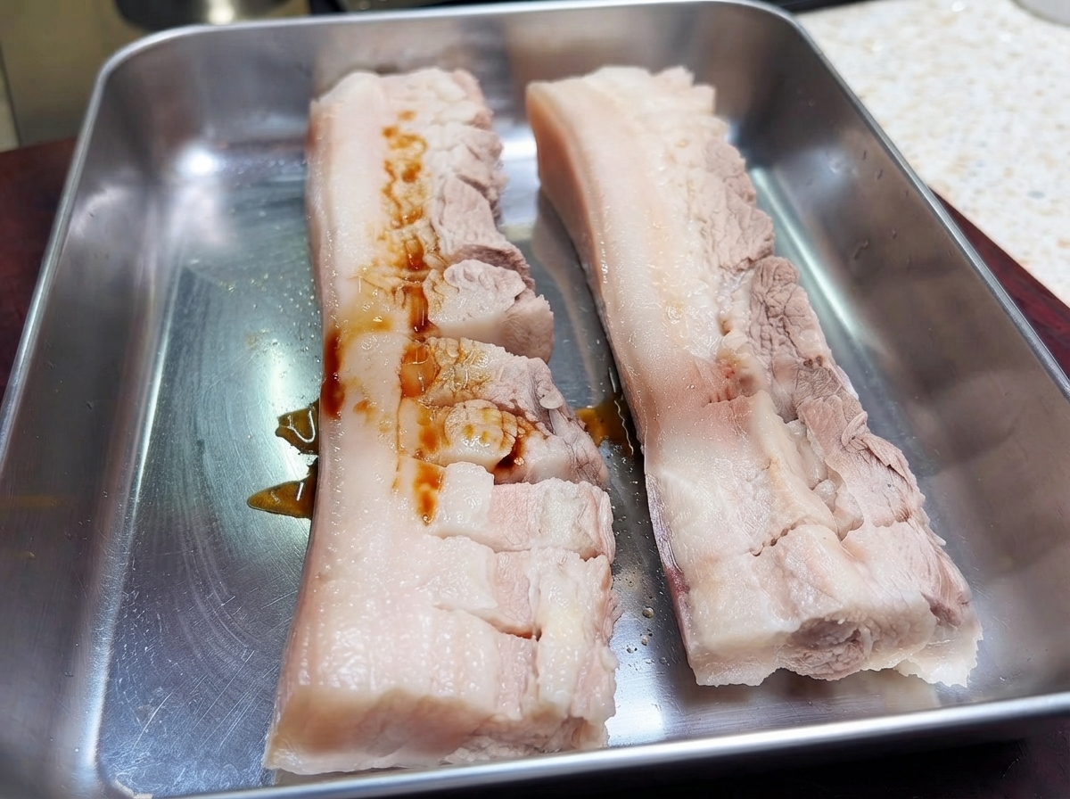 金属製のトレイに置かれた2つの切れ目を入れた豚バラ肉に、濃い醤油が注がれている様子。
