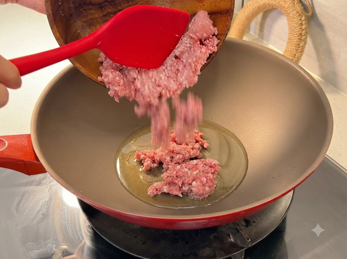 木のボウルから、油を熱した赤い中華鍋に新鮮な豚ひき肉が入れられている様子。