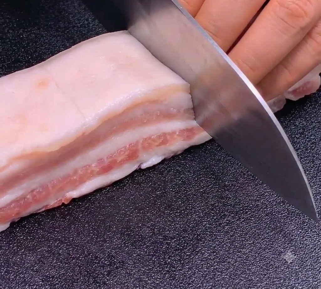 暗い色のまな板の上で、鋭い包丁を使って豚バラ肉に正確で均一な切り込みを入れている様子。