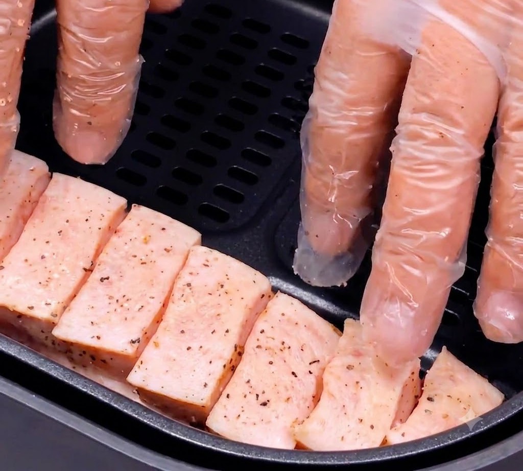 手袋をした手が、味付けした豚バラ肉をエアフライヤーの黒いバスケットの中に丁寧に配置している様子。