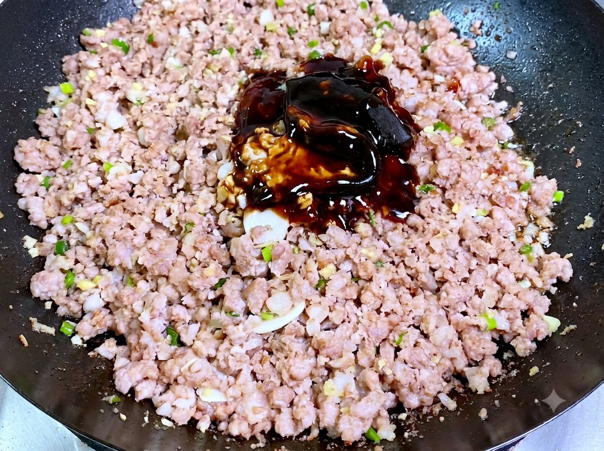 中華鍋の中で、炒めた豚ひき肉の上に濃い色の豆味噌とソースの山が乗せられているところ。