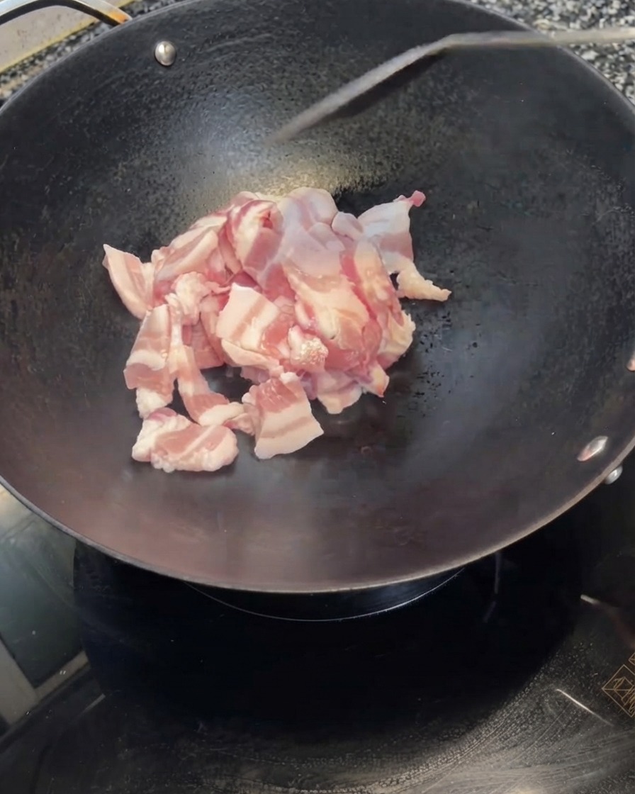 白いボウルからスライスされた豚バラ肉が熱した黒い中華鍋に投入されている様子。
