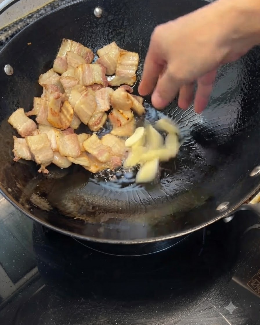 きつね色に焼けた豚バラ肉が入った中華鍋に、潰したニンニクが投げ入れられている様子。