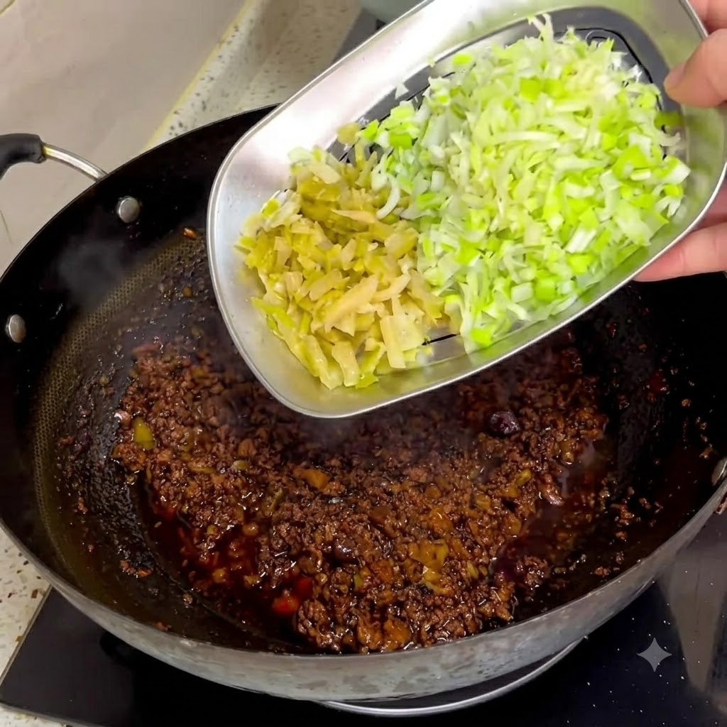 メタルトレイから、中華鍋の中の濃い色の肉味噌へ刻んだザーサイと青ネギを滑り込ませている様子