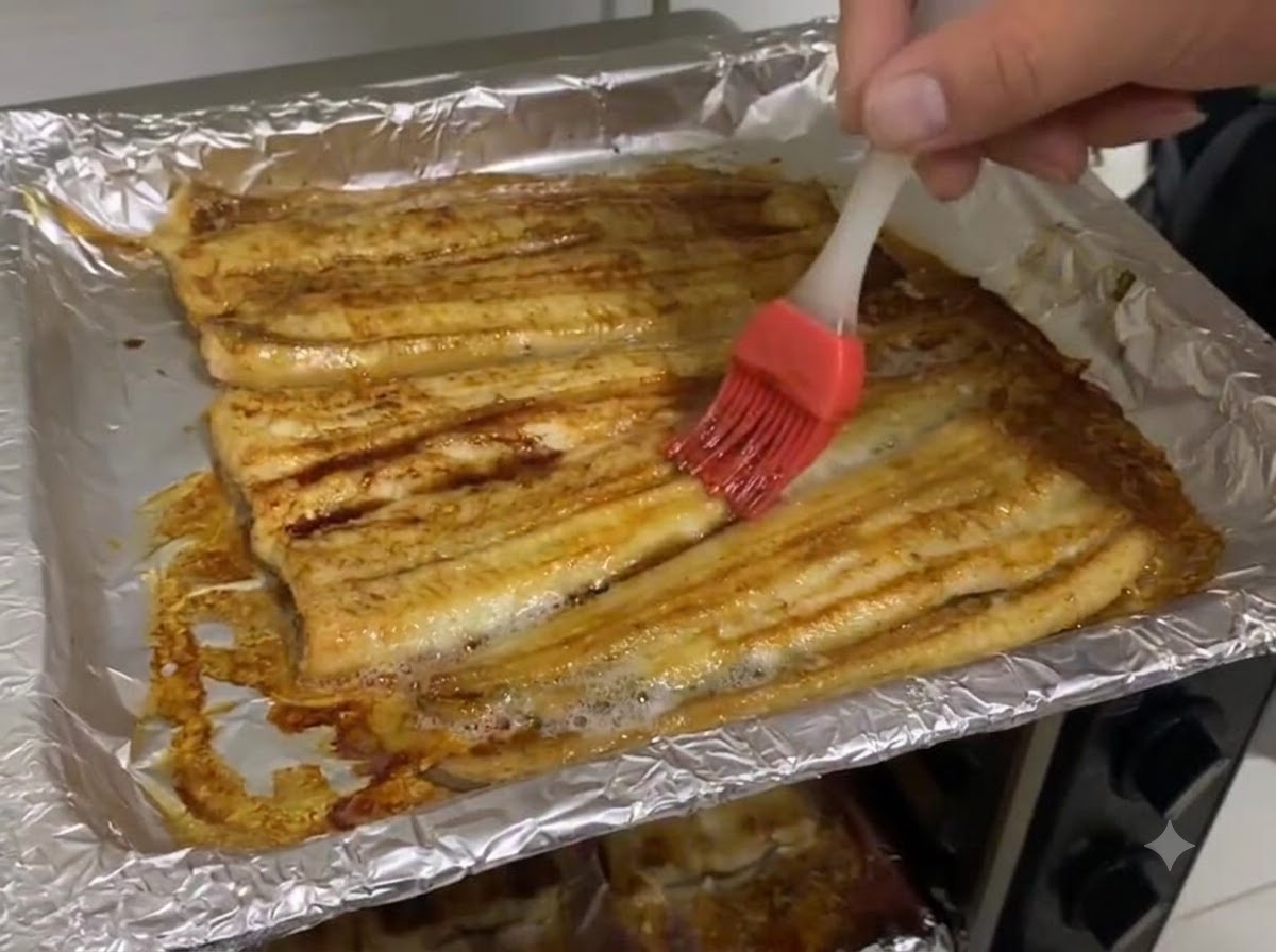 ホイルを敷いた天板の上の焼きうなぎの表面に、濃口醤油を刷毛で塗っているところ。