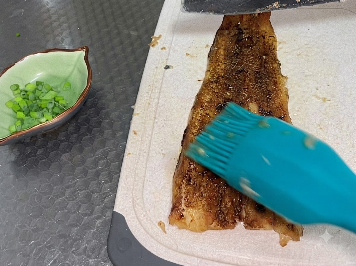 刻みネギが入った緑色の小鉢の隣で、焼き上がったうなぎのフィレを白いまな板の上でスライスしている様子。
