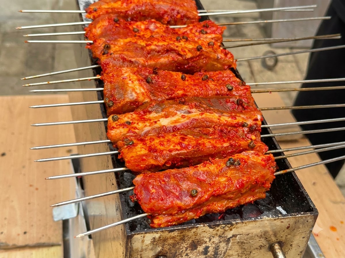 赤い衣をまとった複数の串焼きリブが、炭火の上で並んで焼かれているところ。
