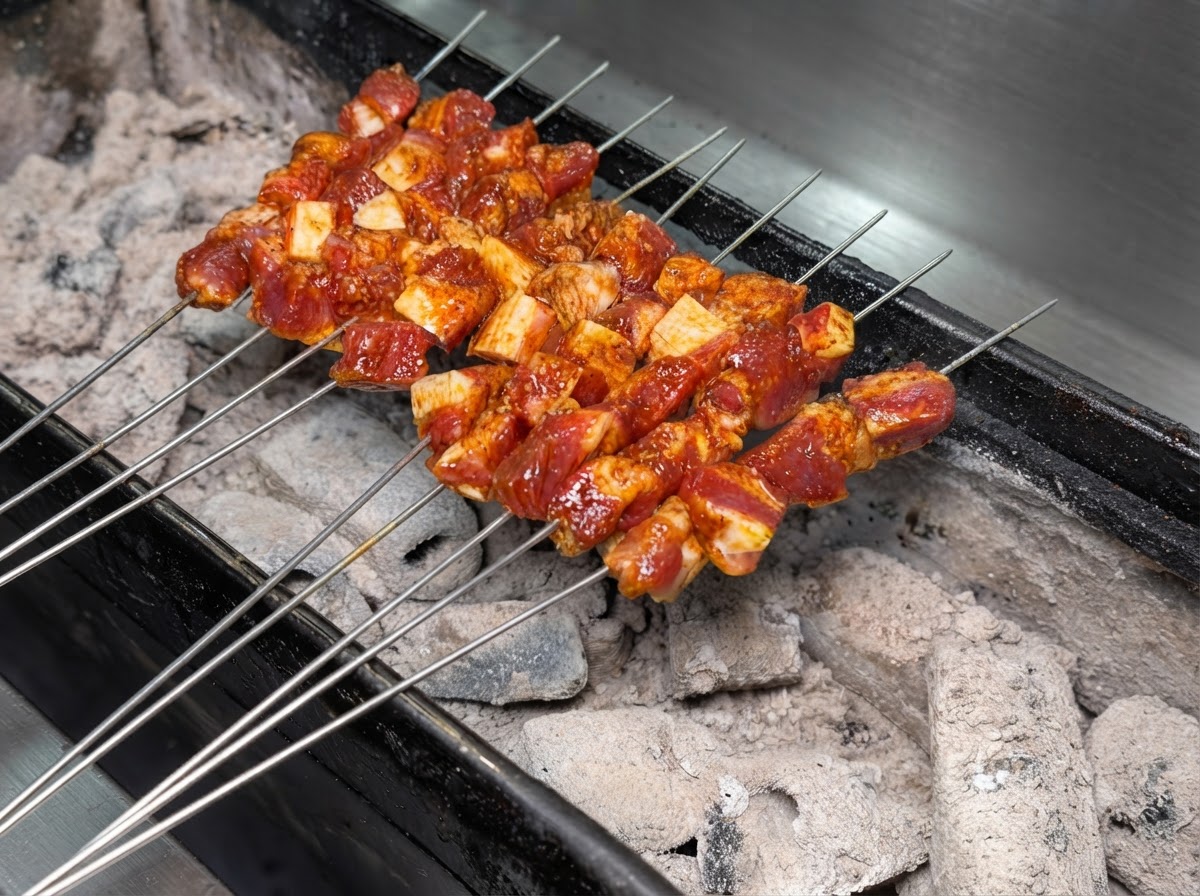 味付けされた豚肉の金属串が、燃え盛る炭火グリルの上にきれいに並べて焼かれている様子。