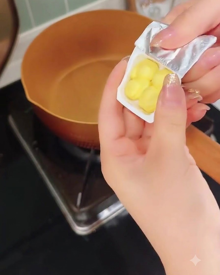 温まった銅色のフライパンの上で、黄色いバターの小片が入った小さなプラスチック容器を持っている手。