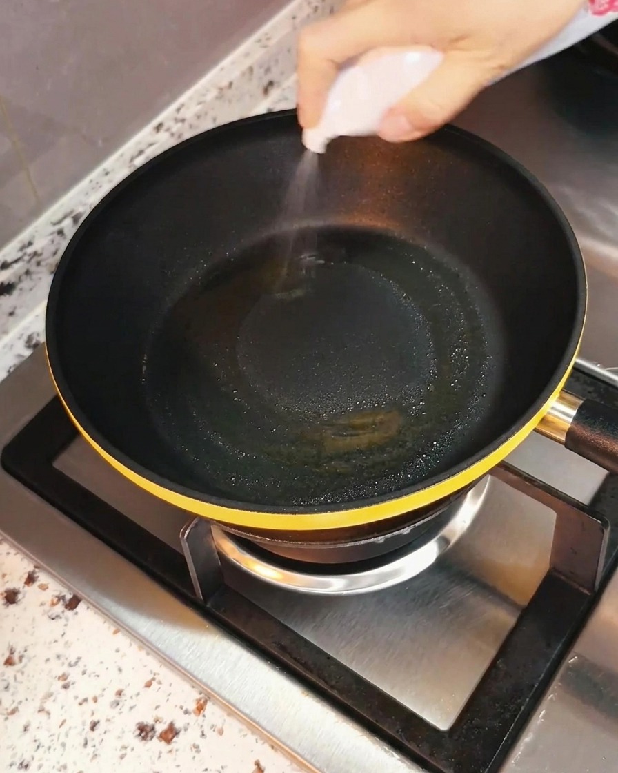 コンロのバーナーに置かれた黒いフライパンの中に、調理油が均等にスプレーされている手。