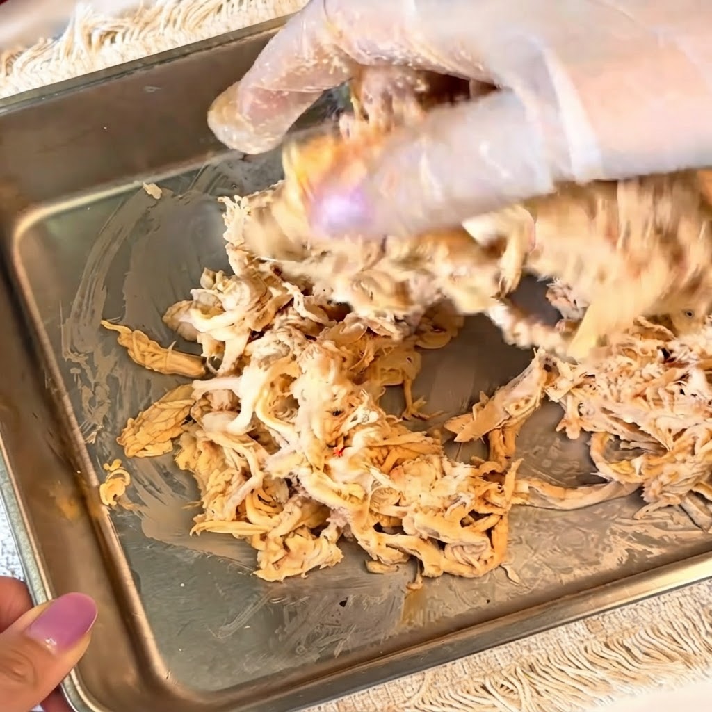 手袋を着用した手が、金属製の容器の中で味付けしたほぐしチキンを混ぜ合わせている様子。