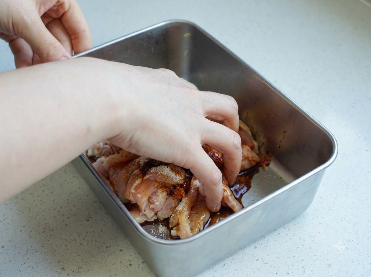 金属製容器の中で、スライスした鶏肉と調味料を手で揉み込んでいる様子。