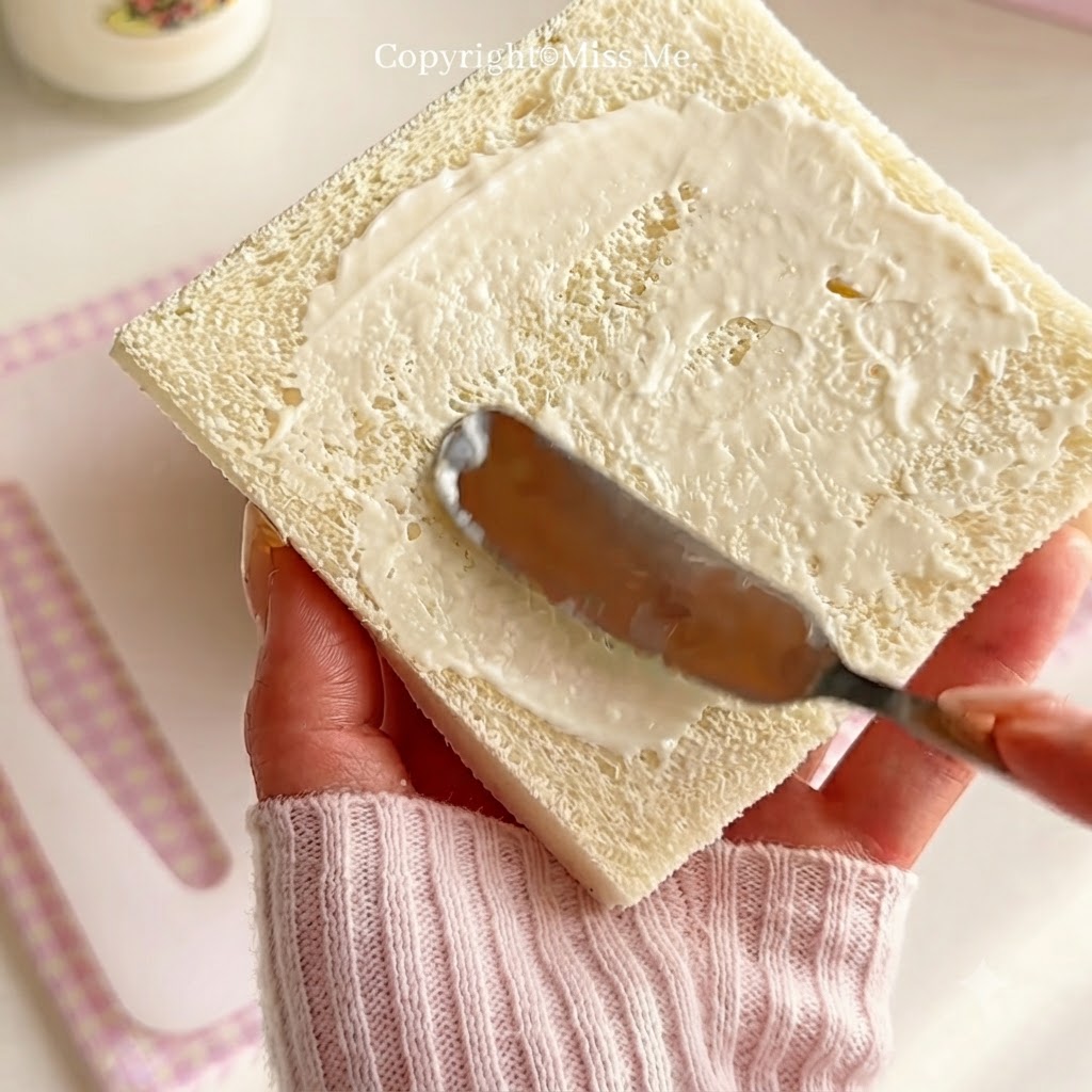 ナイフを持ち、四角い食パンの表面全体に白いサラダドレッシングを均等に塗り広げている手。