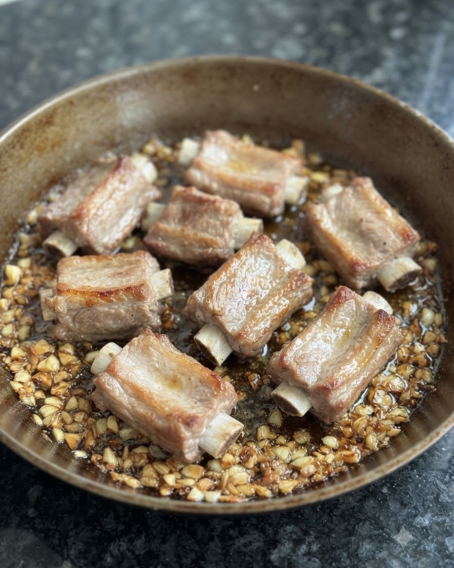 炒めたニンニクとオイルが広がるフライパンで焼き色をつけられている下茹で済みのスペアリブ