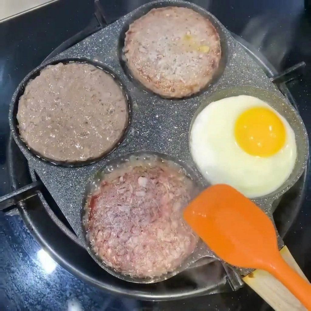 オレンジ色のスパチュラで裏返されるハンバーガーパティと、フライパンの別の区画で焼かれている目玉焼き。