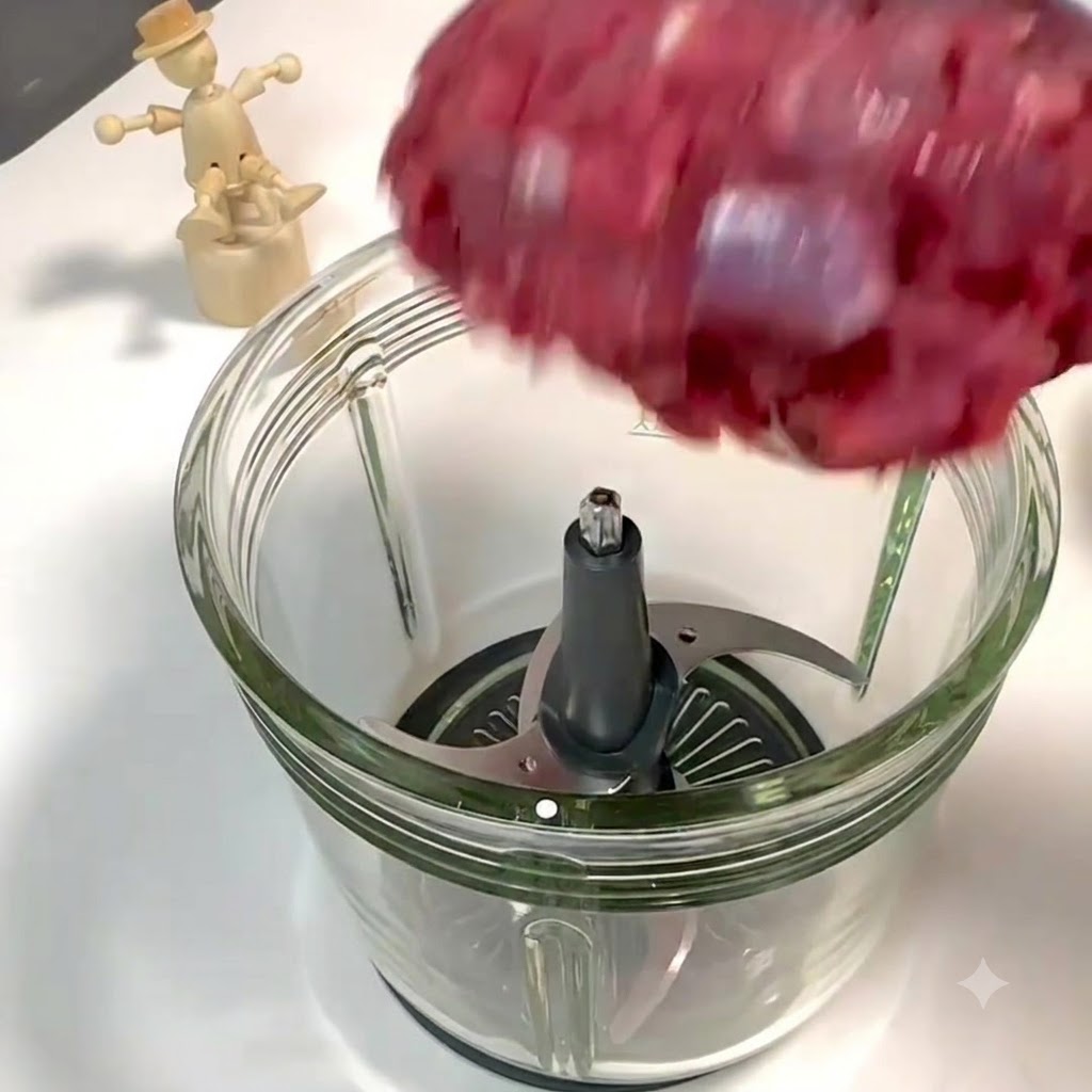 一口大に切った牛もも肉と豚バラ肉が、フードプロセッサーの金属刃の上部から開いたガラス容器に投入されている様子。