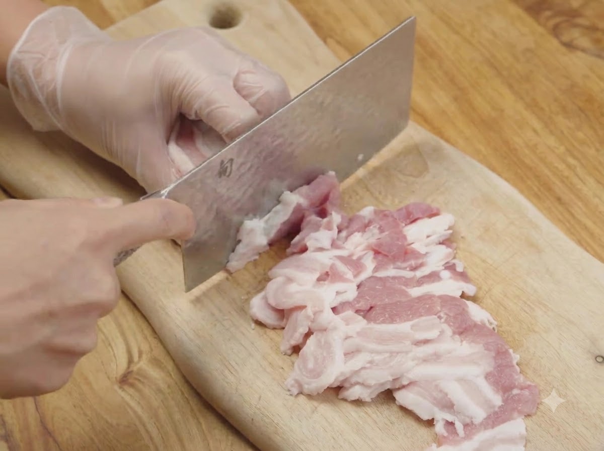 キッチン用の包丁を使い、木のまな板の上で生の豚バラ肉を薄切りにしている様子。
