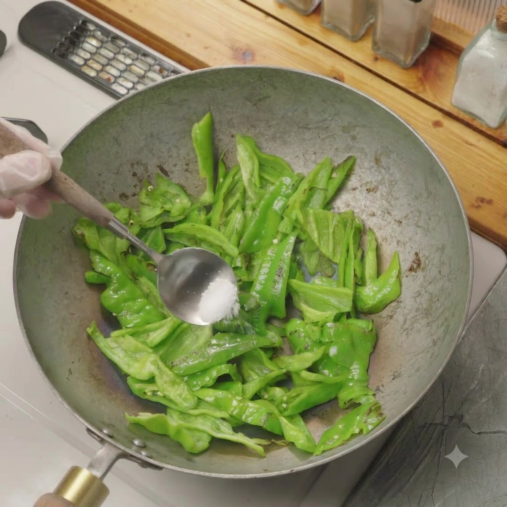 中華鍋で乾煎りされているスライスしたピーマンに、スプーン一杯の塩が加えられている様子。