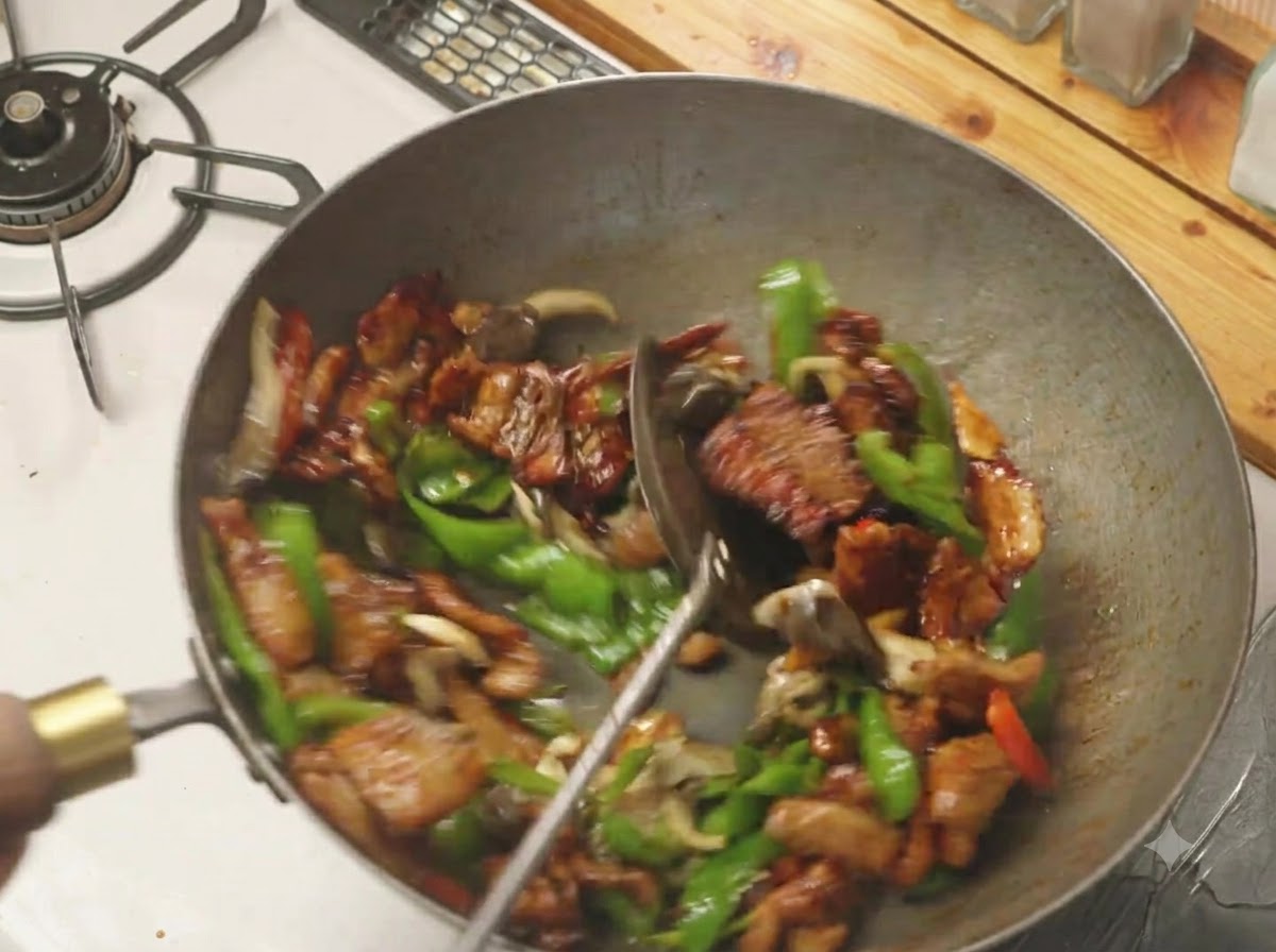 中華鍋の中でピーマン、キノコ、豚バラ肉がヘラを使って手際よく混ぜ合わされている様子。