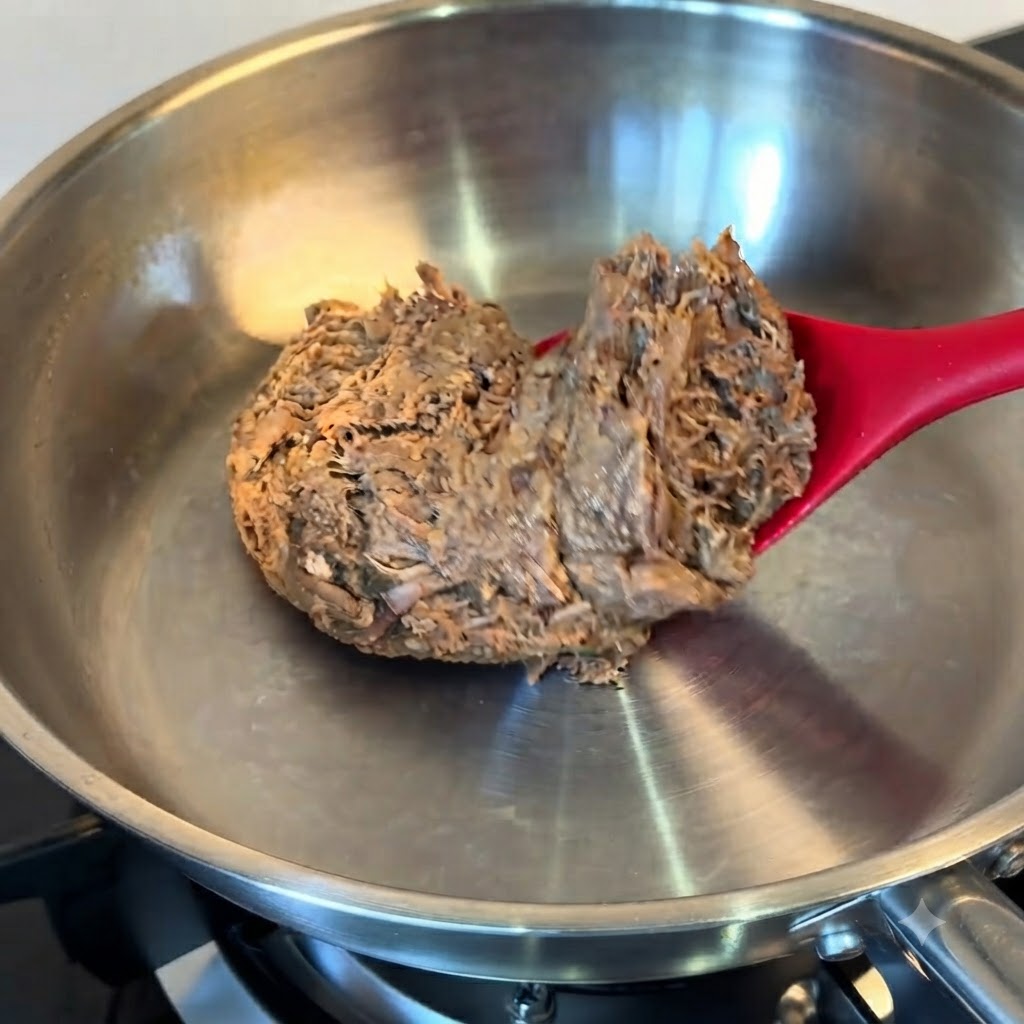 赤いスパチュラを使い、解凍したほぐし牛肉の塊をステンレス製のフライパンに入れている様子。