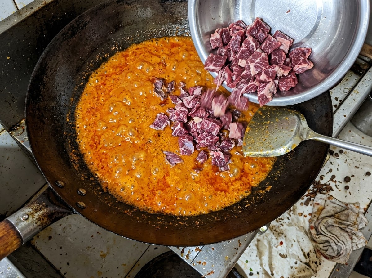 金属製ボウルから、泡立つ赤色のラー油が入った中華鍋に牛バラ肉の塊が入れられている様子。