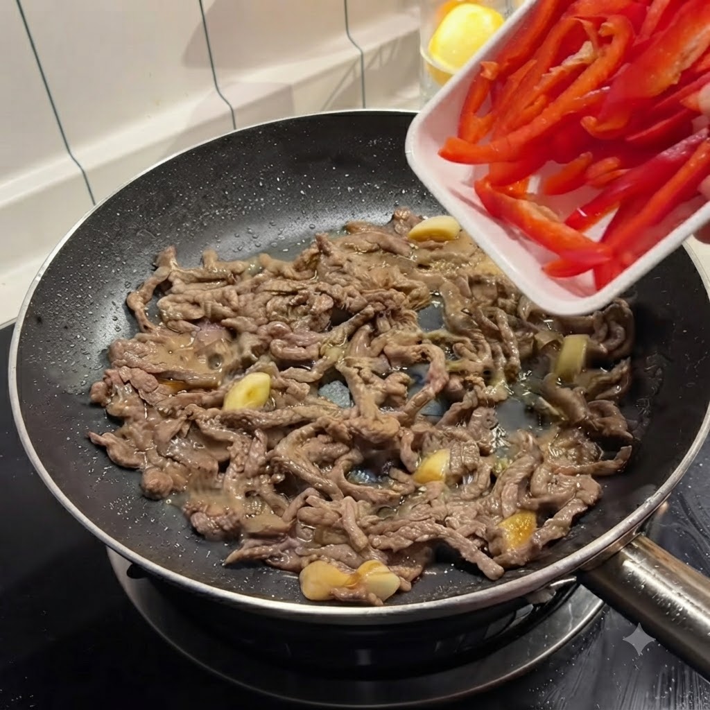 焼き色のついた牛肉が入ったフライパンに、白いトレイから赤パプリカを流し入れている様子。