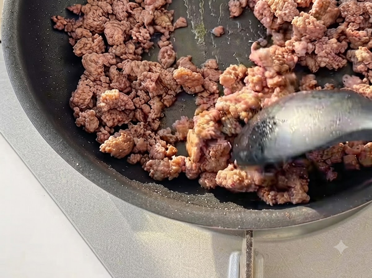 黒いフライパンの中で、スパチュラを使って牛ひき肉をきつね色になるまで炒めている様子
