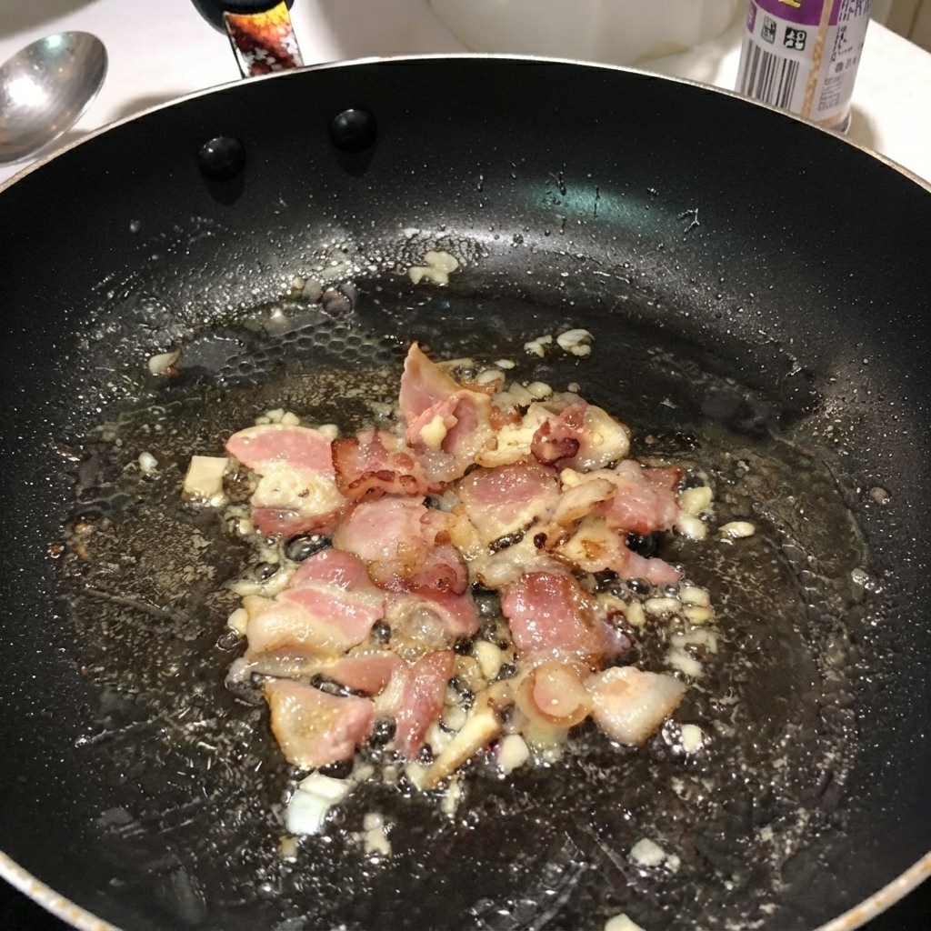 黒いフライパンの中で、茹でたペンネパスタがベーコンや玉ねぎと混ざり合っている様子。