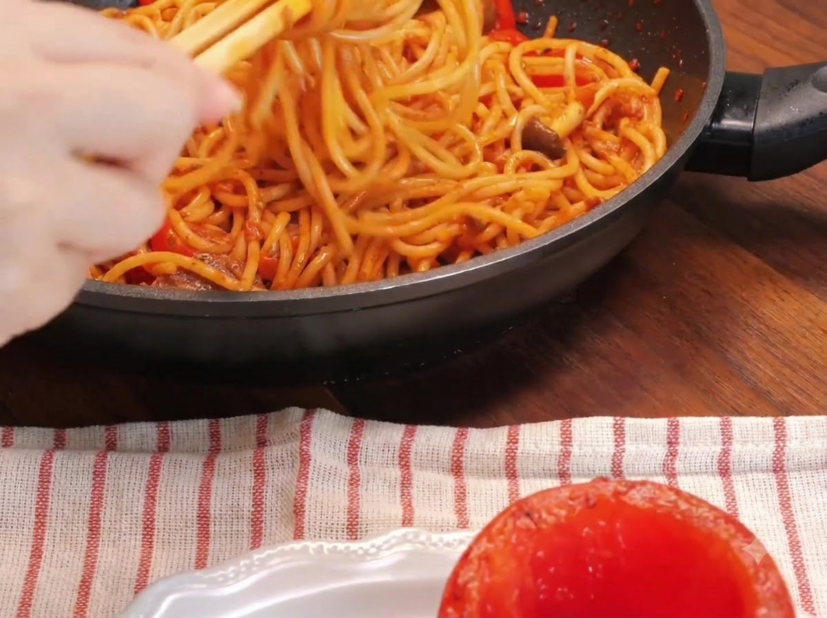 フライパンの中でスパゲッティを箸で巻き取り、皿に置かれたローストトマトの器に盛り付けている手元のクローズアップ。