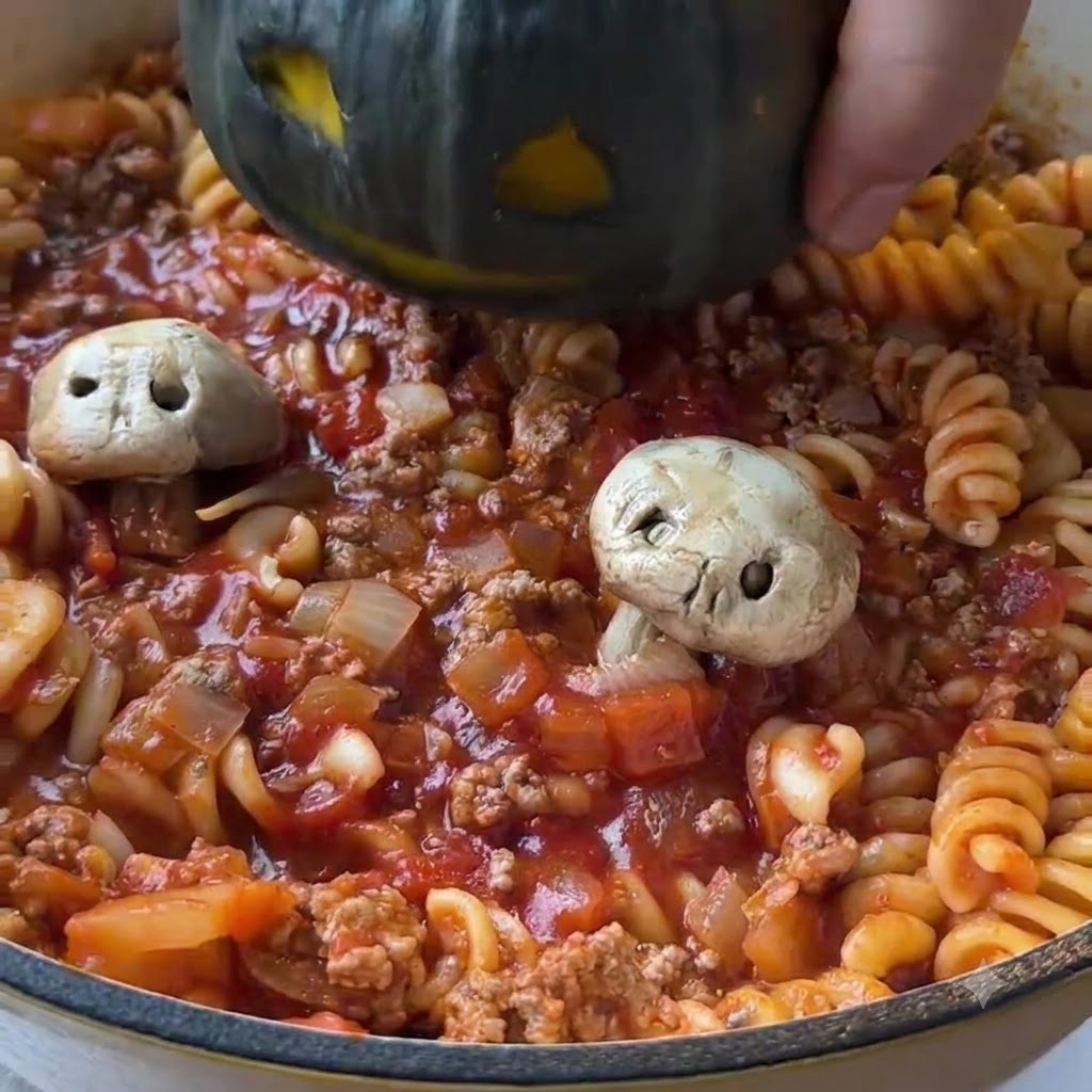 顔が彫られた濃い緑色のミニカボチャが、味付けされたトマトパスタでいっぱいのフライパンの中心に下ろされているところ。