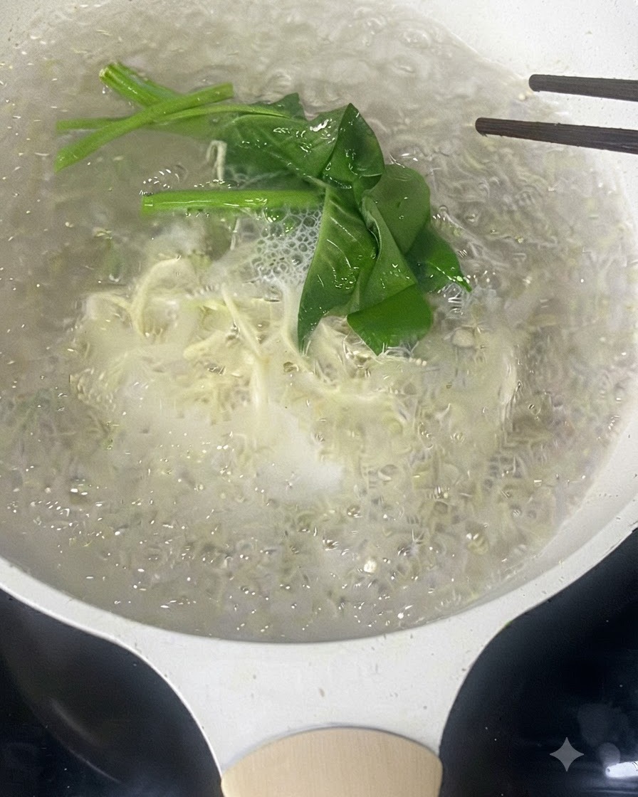 箸を使って、細い白い麺と緑の葉物野菜が沸騰したお湯の中で一緒に混ぜられている様子。