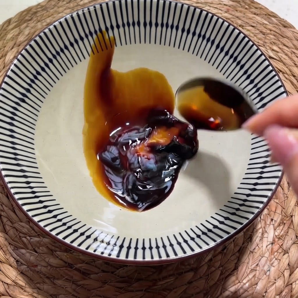 柄のある陶器のボウルに入ったオイスターソースに、スプーンから濃口醤油を注いでいる手。