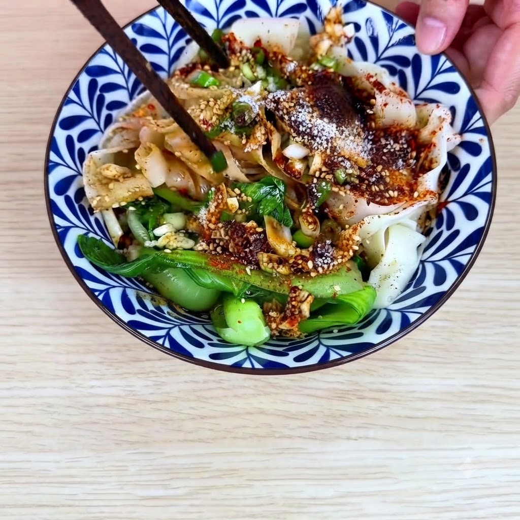 陶器のボウルの中で、平打ち麺、チンゲン菜、チリソースを箸で混ぜ合わせている様子。